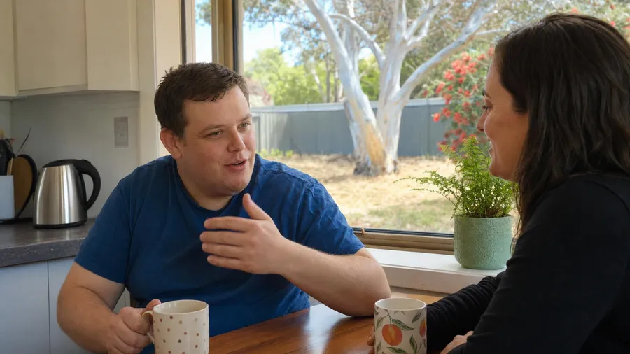 A man holding a mug and talking with a support worker at a sunny kitchen table