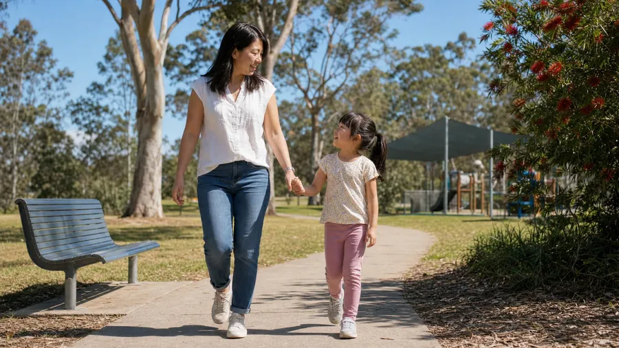A mother and her young daughter walking hand in hand along a park path