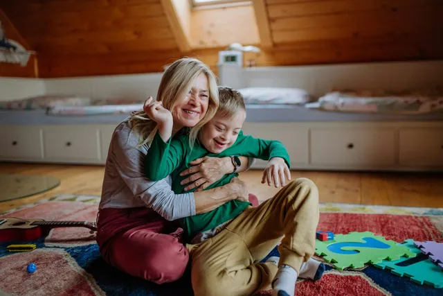 A woman and a young boy laughing and hugging on a colourful rug at home