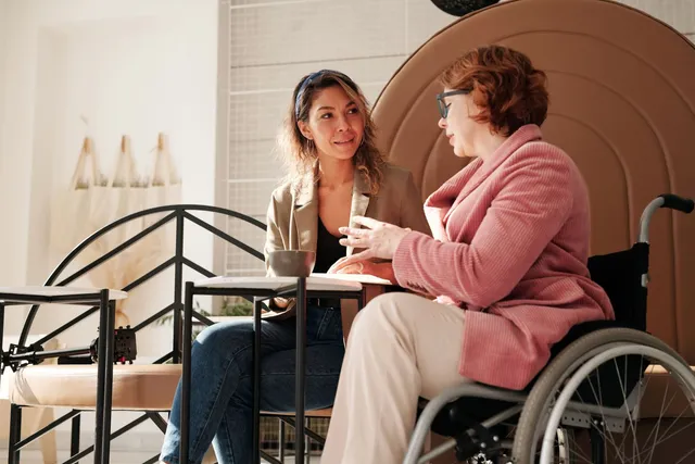 Two women talking over a table in a cafe, one using a wheelchair, in a supportive conversation