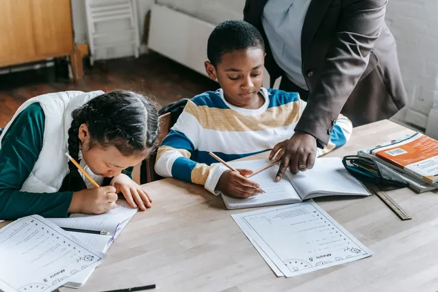 A teacher standing beside two young students writing at a classroom desk
