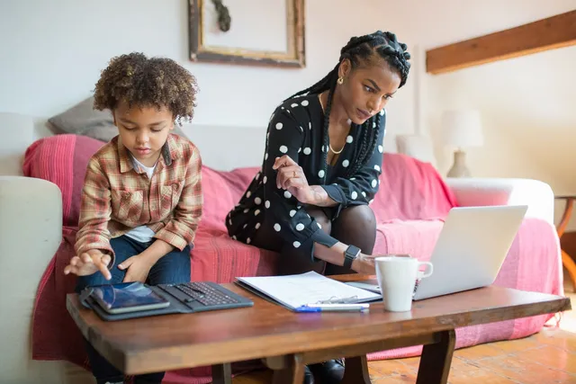 A mother and her son at a wooden table with a laptop and papers while she makes notes