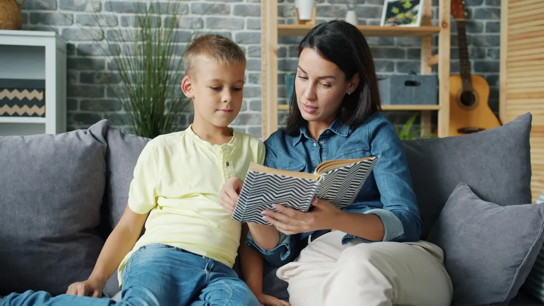 A mother and her son sitting on a sofa reading a book together