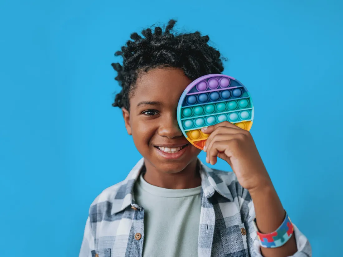 A smiling child holding a colourful pop-it toy over one eye against a blue background
