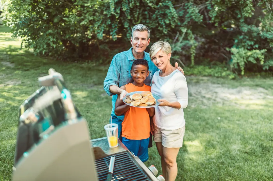 A man, a woman and a boy holding a plate of food around a barbecue in a backyard