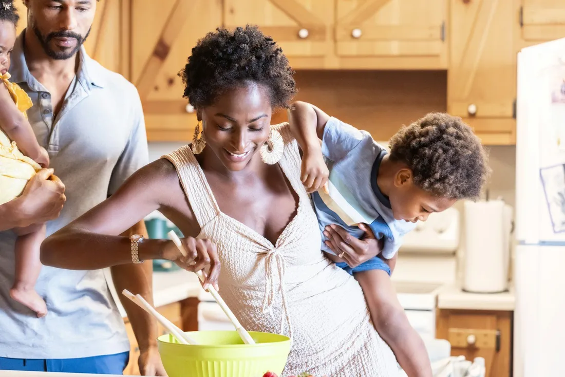 A mother cooking in the kitchen while her young son rides on her back and a man looks on