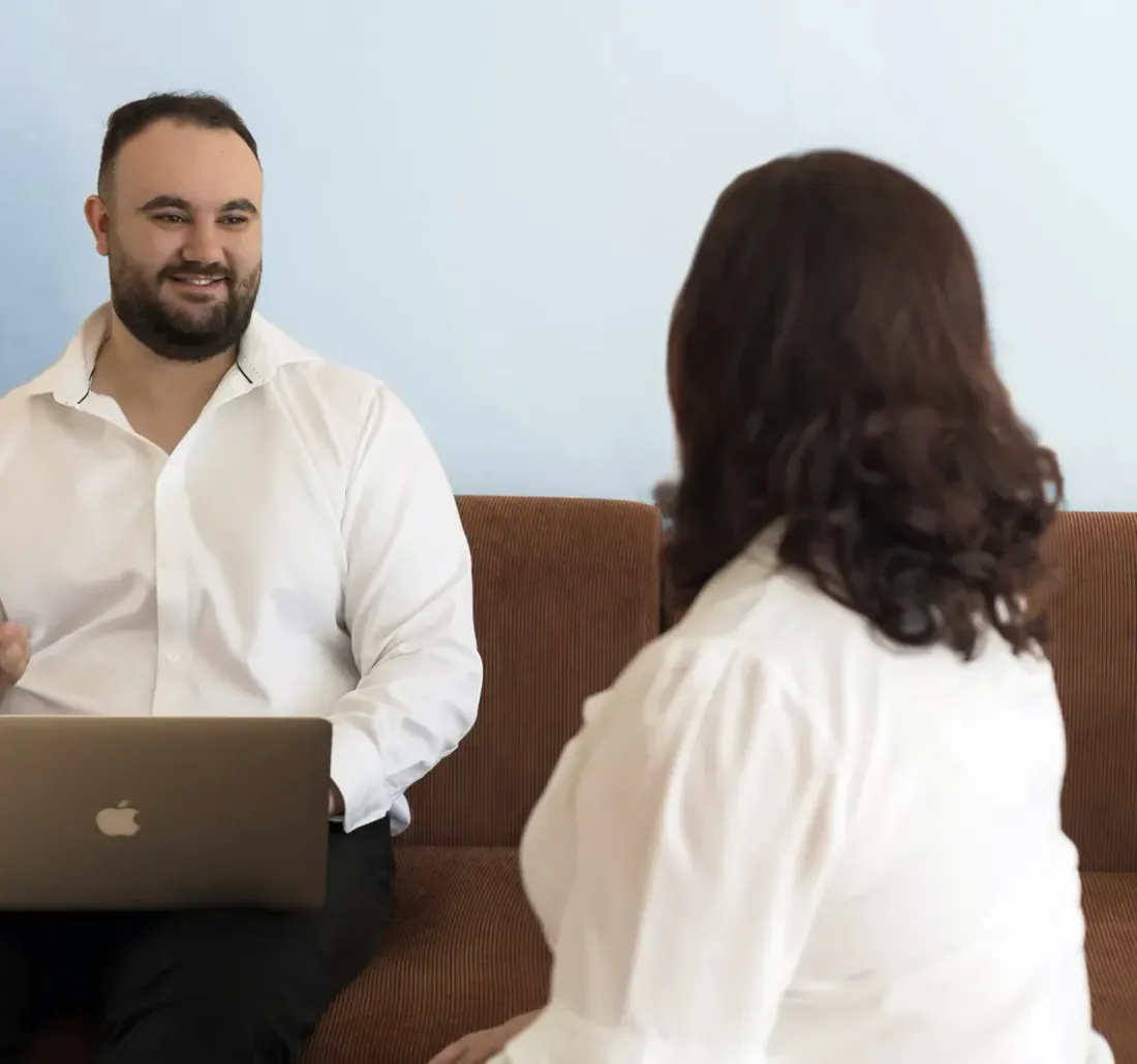 A smiling man in a white shirt with a laptop talking with a woman on a sofa