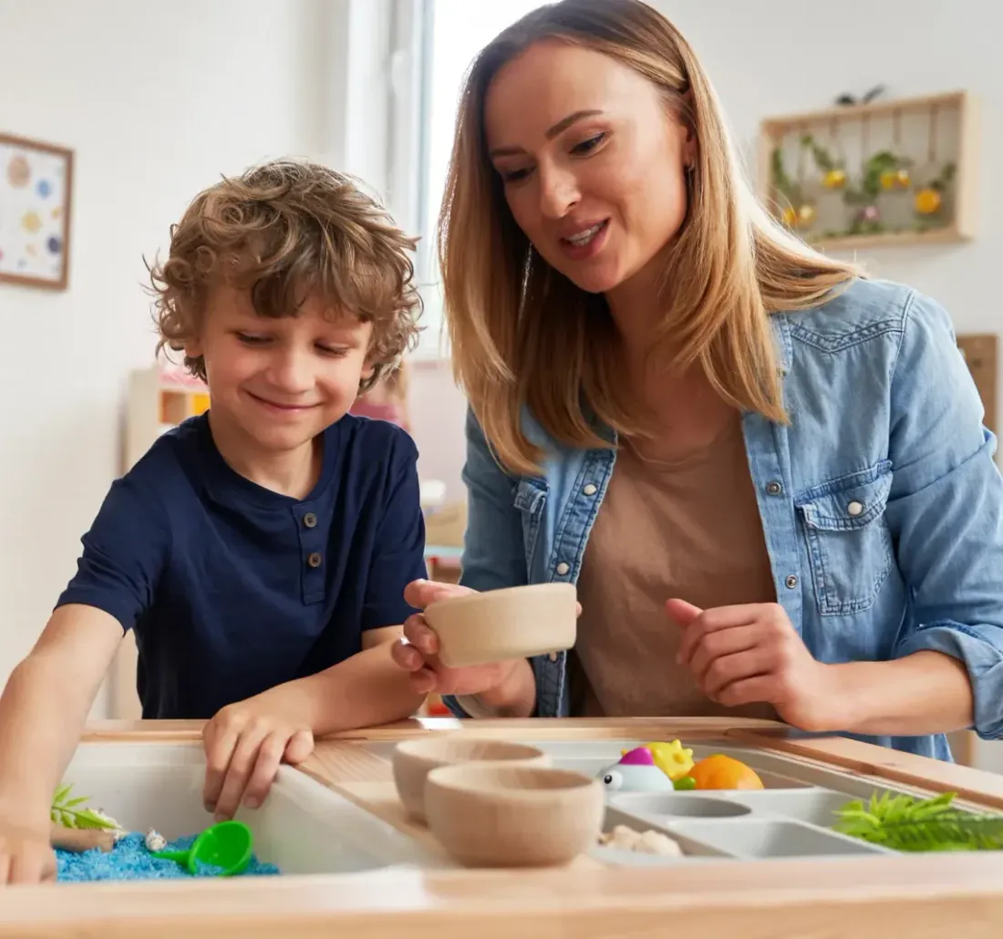 A mother and her young son exploring a tray of sensory play materials at a table
