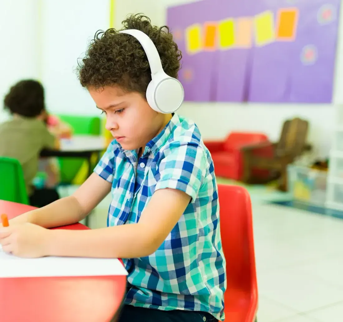 A boy wearing headphones drawing at a table in a bright classroom