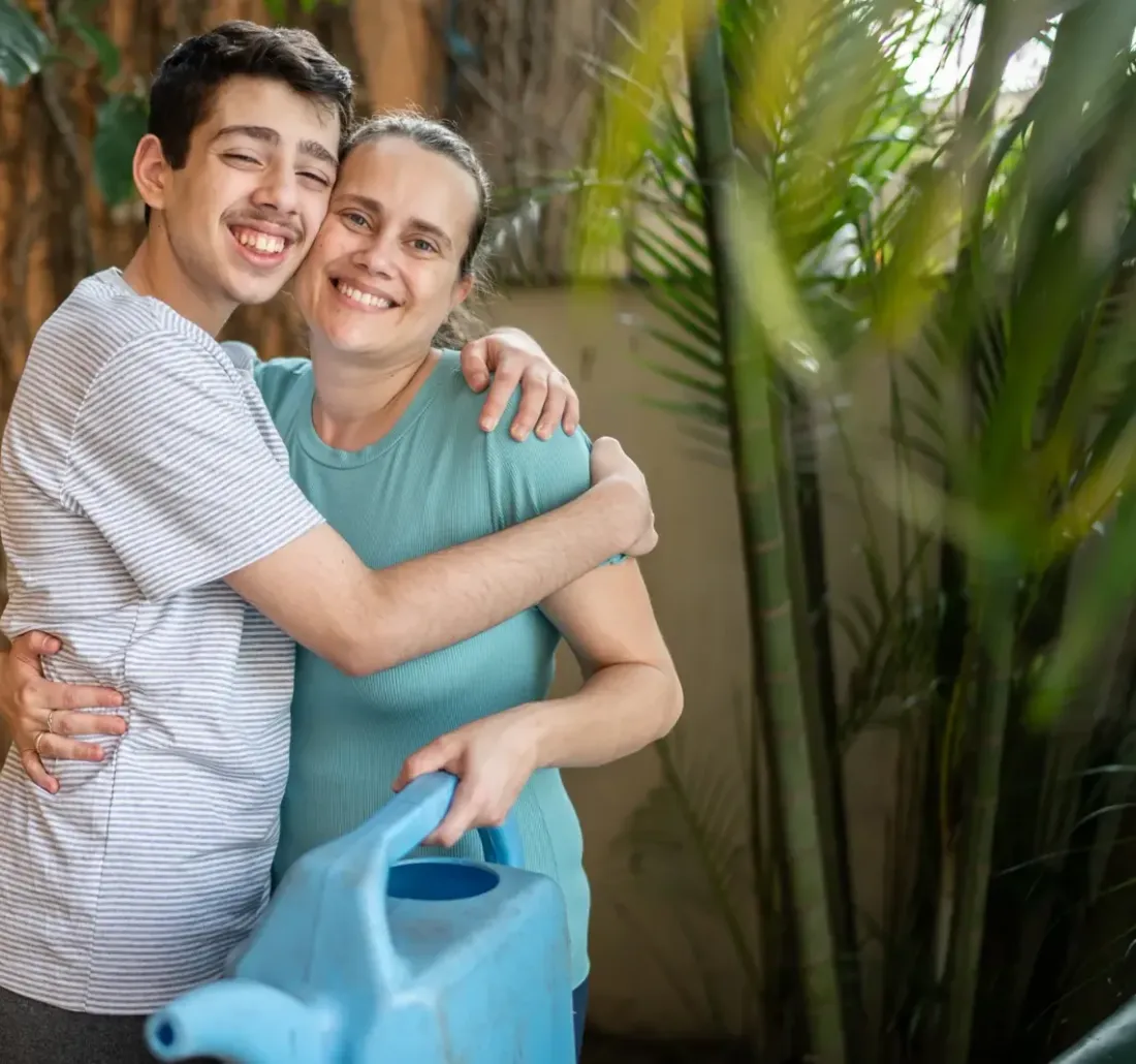 A teenage boy hugging his mother in a garden, holding a blue watering can