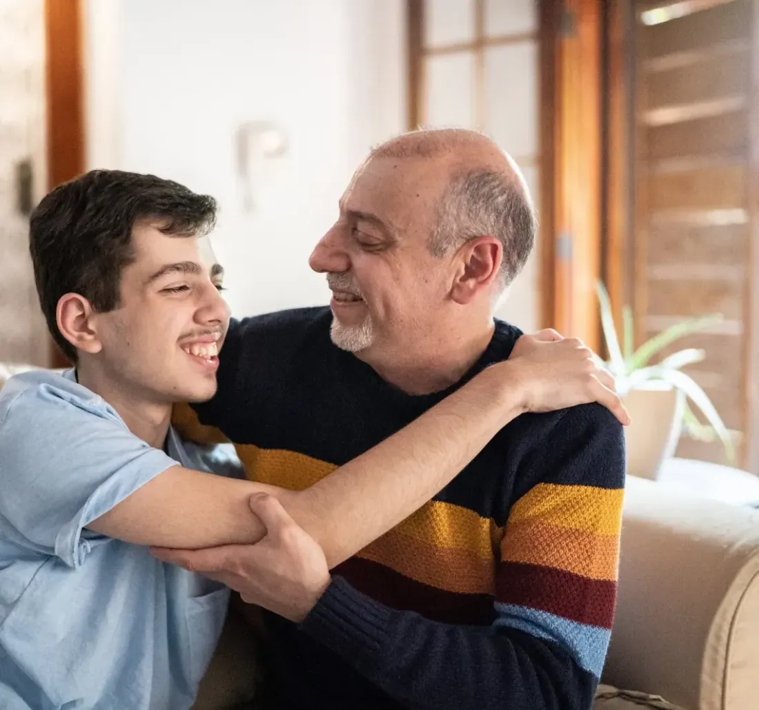 An older man and a teenage boy sharing a hug and a laugh on a sofa at home