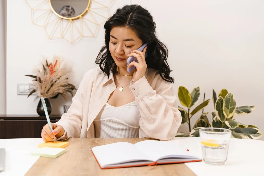 A woman on the phone writing notes at a table in a bright room