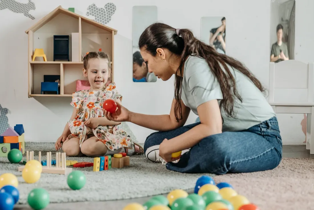 A woman sitting on the floor playing with a young girl and colourful balls and blocks